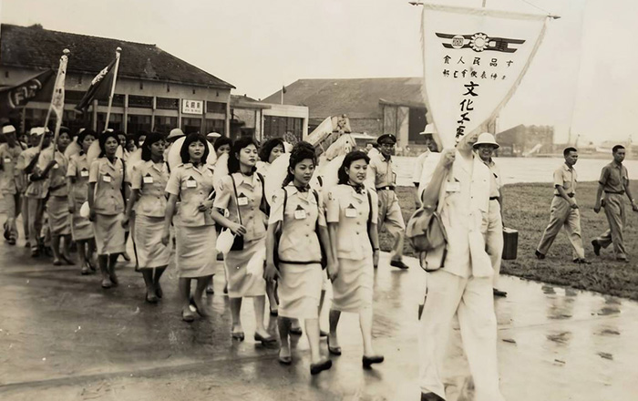 老照片為民國 46 年端午節前夕，電信勞軍團於台北松山機場整隊出發到金門的紀念照，隊伍後方有「電信徽大旗」壓陣，穿著整齊制服的電信局女員工，隨行將粽子等端午物資送到金門前線將士的手上。在通訊不便的台灣戒嚴時期，電信人以實際行動，溫暖串連起「軍民一家」的情感。